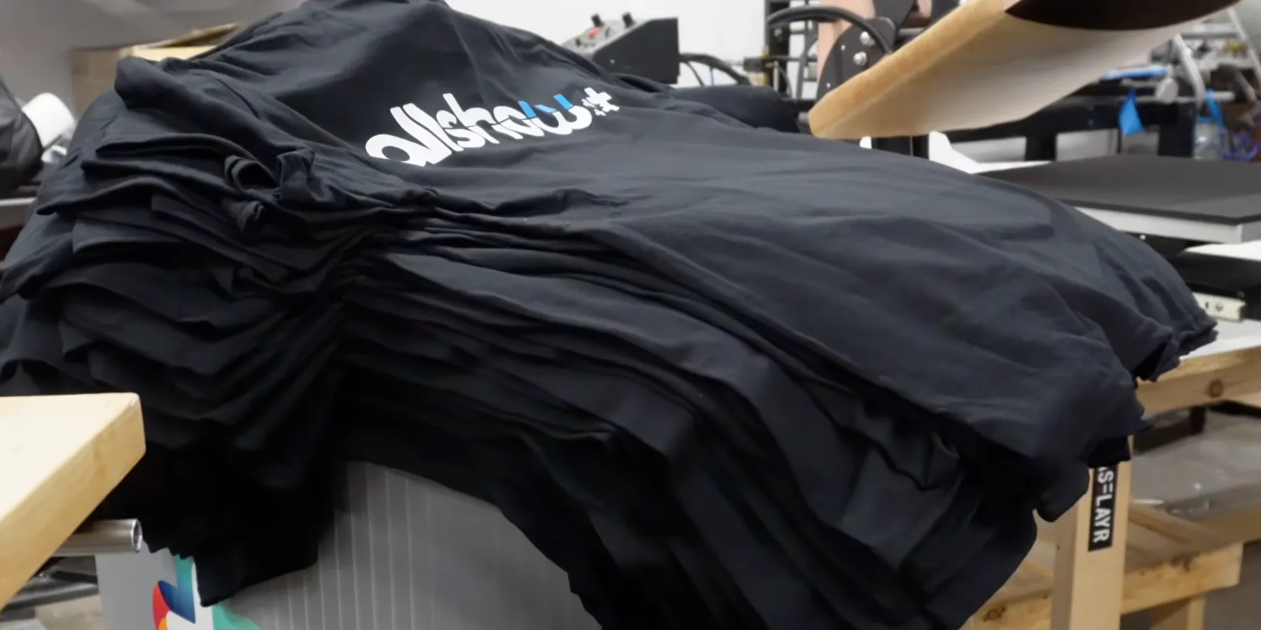 Stack of black Gildan DryBlend shirts with DTF prints ready on an AMS production table near the heat press